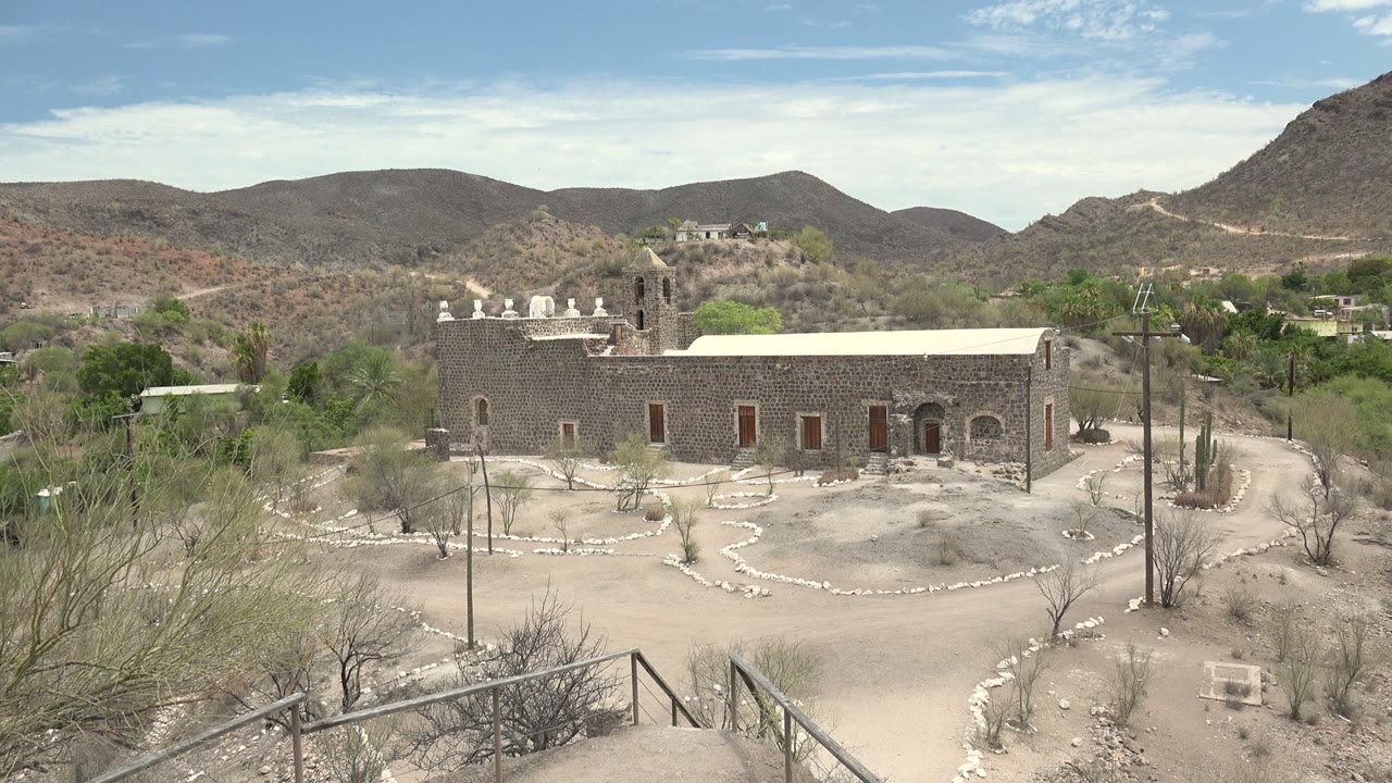 A broad Mulege mission view includes foreground metal railings, pale dirt paths, sparse shrubs, a stone facade, distant homes or structures on the hillside, and no visible people.