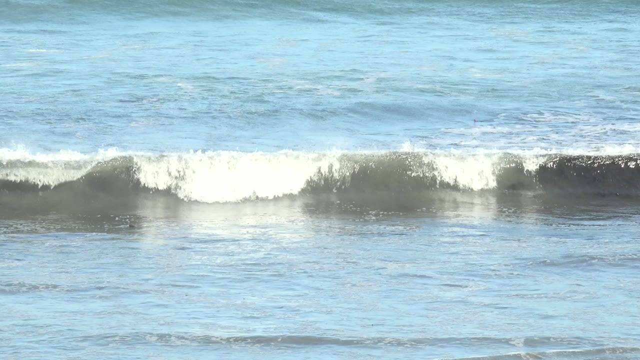 At 0:02 in La Bocana, Baja California Sur, layered blue water and white surf stretch across the frame, with a brighter foam band in the foreground and dark rocks at the bottom, with no visible people.