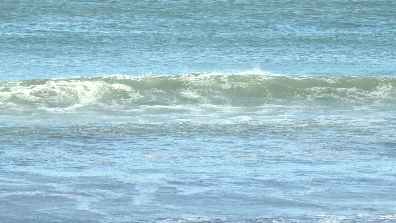 At La Bocana, Baja California Sur, blue-green ocean fills the frame as a low wave line forms with white foam across the upper band, with no visible people.