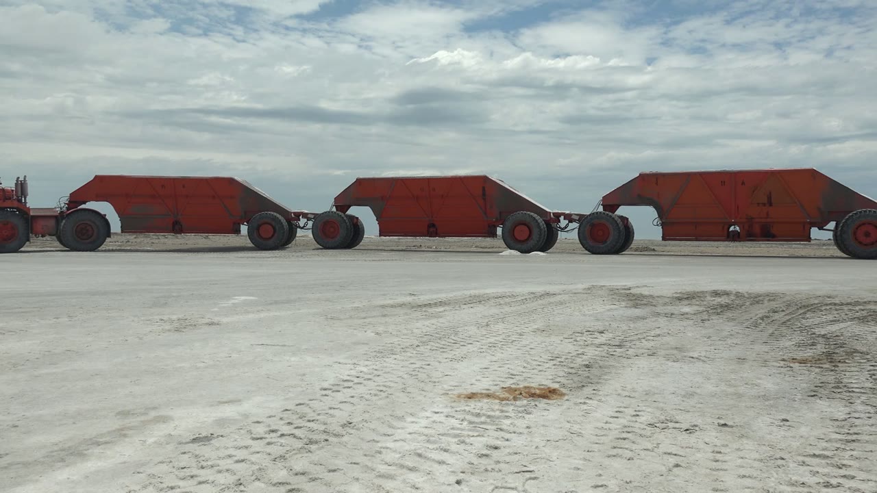 At 0:00, a line of red mine carts moves across pale sandy ground in Guerrero Negro, Baja California Sur, with tire tracks in the foreground and no visible people.
