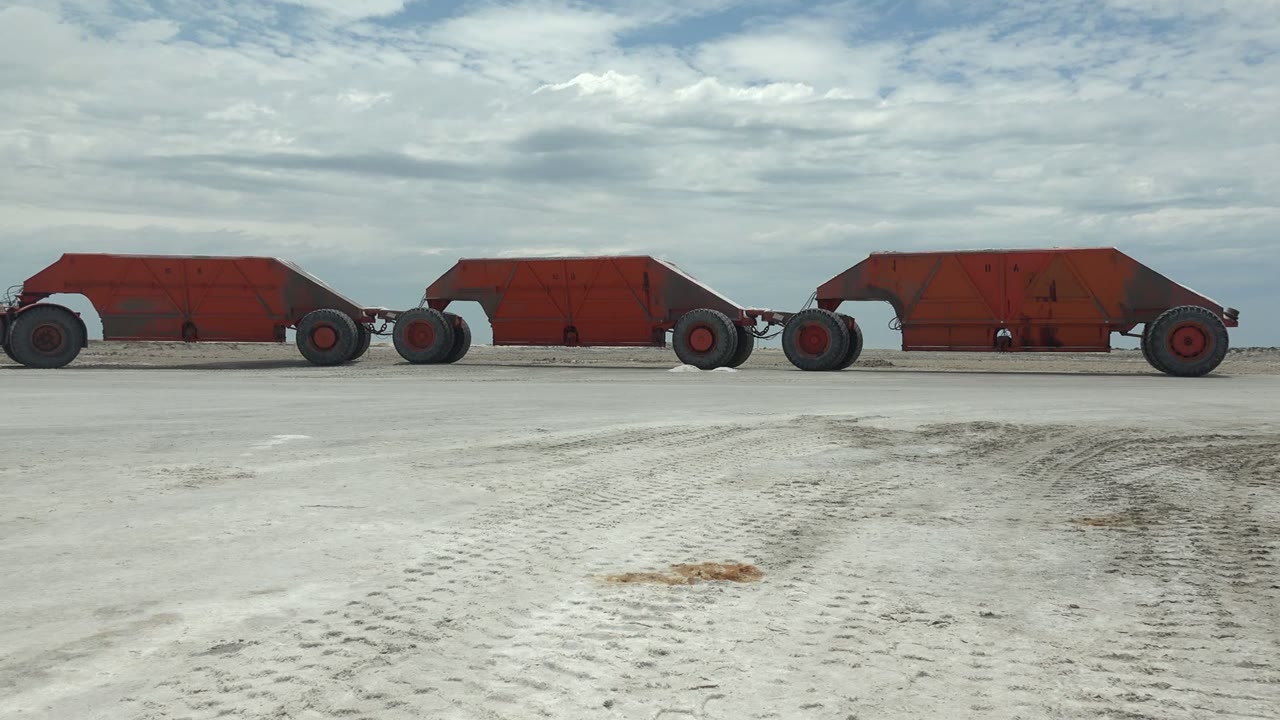 At 0:01, the red carts continue rightward through Guerrero Negro, with the left edge showing part of a vehicle and the sandy foreground marked by wheel tracks, no visible people.