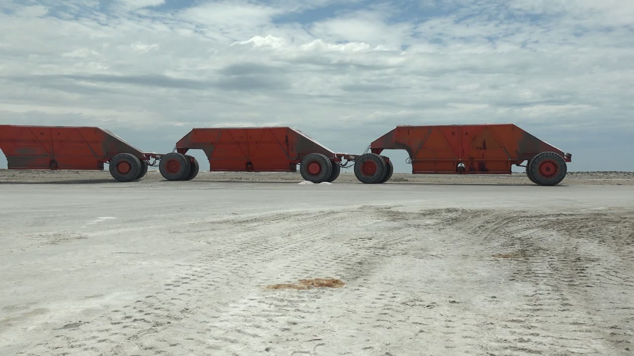 At 0:02, multiple red mine carts span the midground in Guerrero Negro while pale tracked sand fills the foreground and a layered cloudy sky sits above, no visible people.