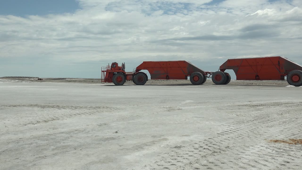 At 0:00 in Guerrero Negro, Baja California Sur, a red hauler pulls red trailers across a broad white flat with a cloudy sky and no visible people.