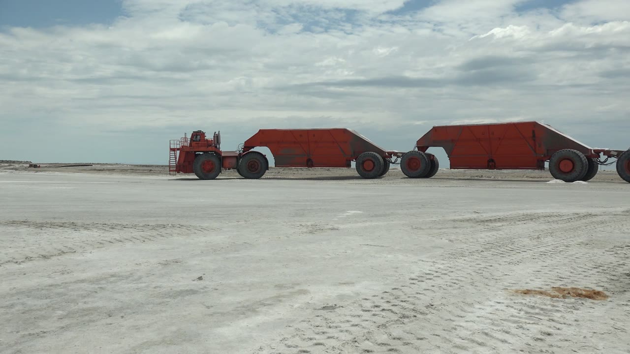 At 0:02 in Guerrero Negro, Baja California Sur, the red hauler and connected red trailers run horizontally across the midground, with empty white terrain in front and no visible people.