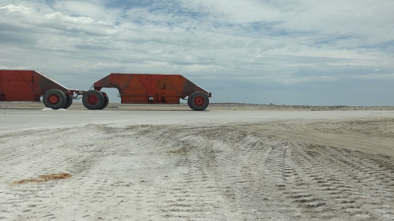 At 0:02, the rusted red vehicle appears farther right in the wide Guerrero Negro frame, with pale dusty ground, track patterns, and no visible people.