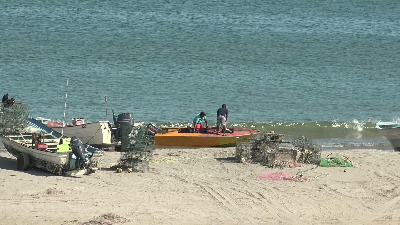 Wide view of San Juanico, Baja California Sur beach with small boats, wire traps, green netting, and two people near an orange boat as light surf reaches the shore.