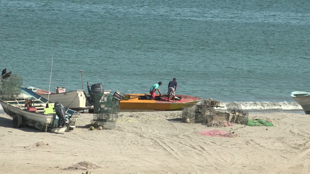 At San Juanico, two people remain near the orange boat, with several beached small boats, wire traps, green netting, and white surf visible against the blue ocean.