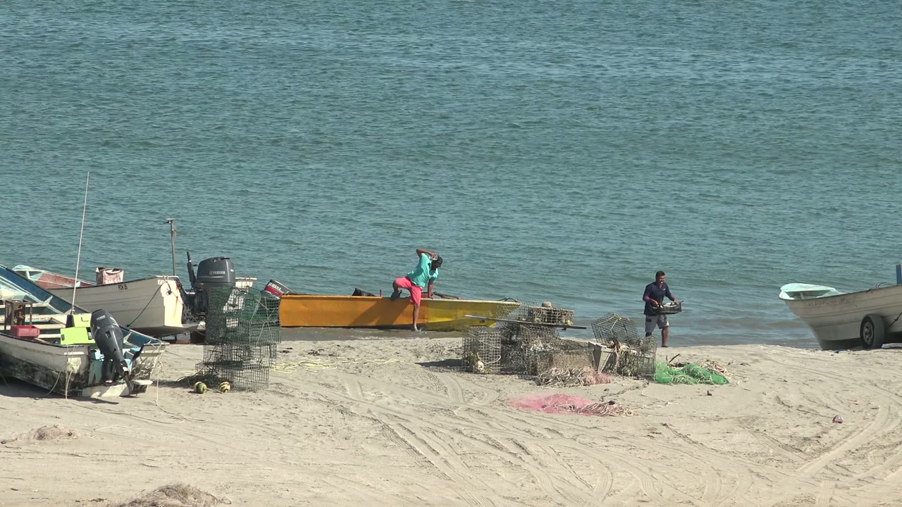 San Juanico, Baja California Sur beach with several small boats on sand, fishing nets in the foreground, one person by a yellow boat, and another person near the surf.
