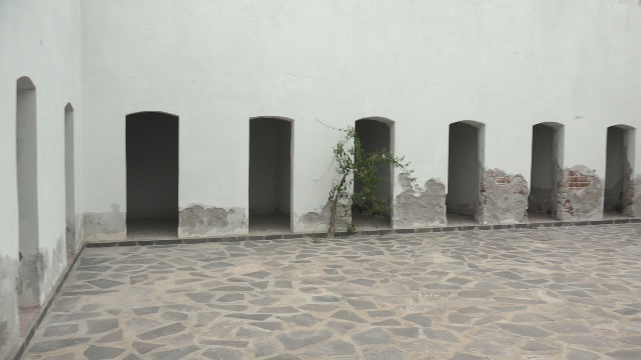 Mulege BCS Mexico stone courtyard with white plaster walls, several arched openings, exposed masonry near the base, a small plant by one opening, and no visible people.