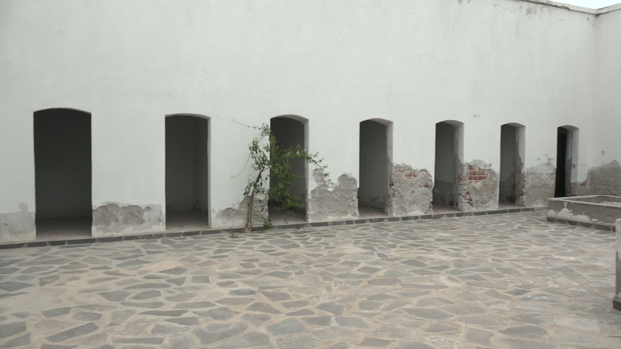Mulege BCS Mexico courtyard with repeated arched wall openings, a low concrete structure entering from the right, weathered plaster, stone paving, and no visible people.