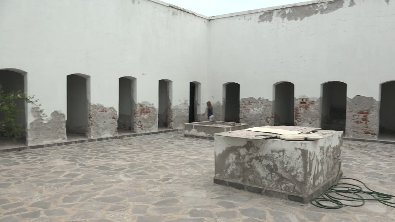 A Mulege, Baja California Sur courtyard appears in a wide view with a person near the left arched openings, a worn stone structure in front, and a green hose on the paving.