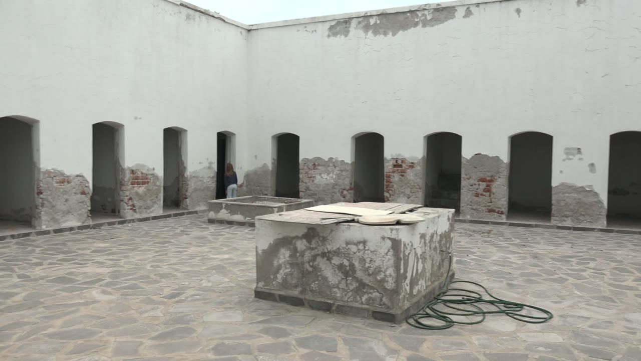 A wide static frame shows the person near the center-left arches of the Mulege courtyard, with patched white walls, a rectangular stone structure, and coiled green hose in front.