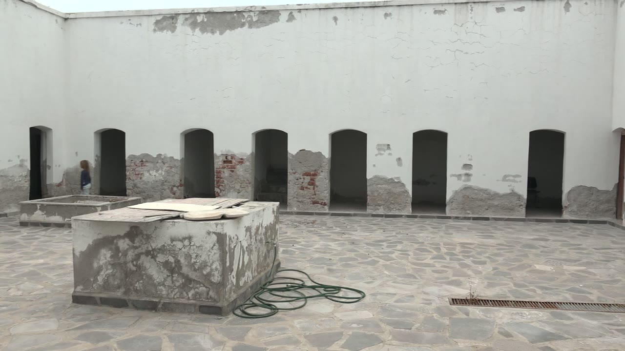Mulege, Baja California Sur courtyard with a weathered white wall, several dark arched openings, a stone table, green hose, and a person briefly visible near the far-left arch.