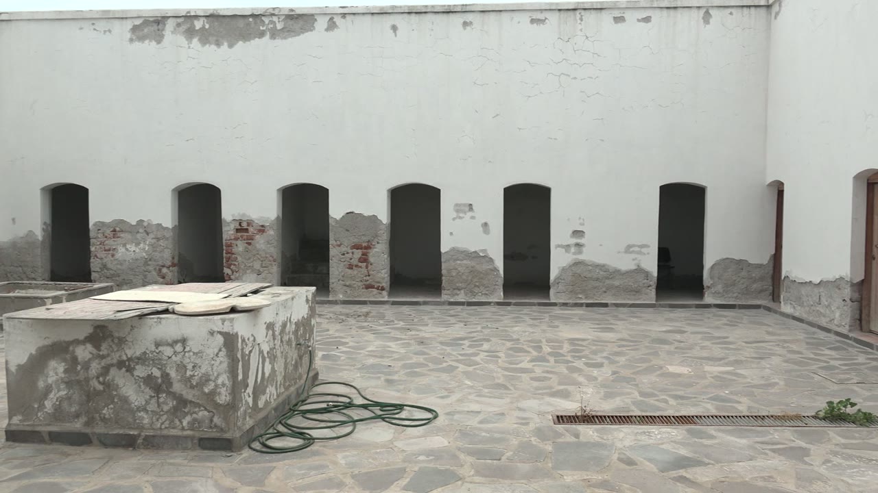 Mulege, Baja California Sur courtyard with a centered stone table, coiled green hose, gray cobblestone paving, peeling white plaster, and no visible people.