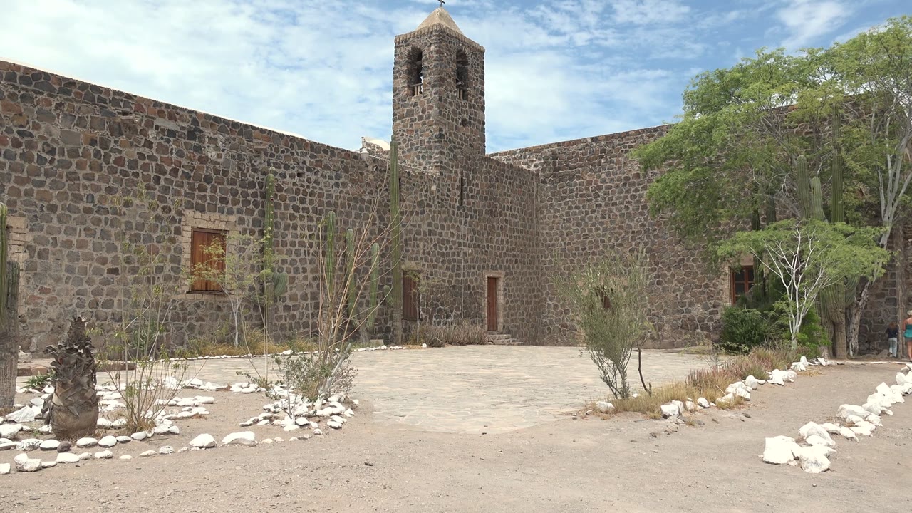 Opening frame of a stone mission courtyard in Mulege, Baja California Sur, with a bell tower rising above dark stone walls, cacti and white rocks in the foreground, and no visible people.