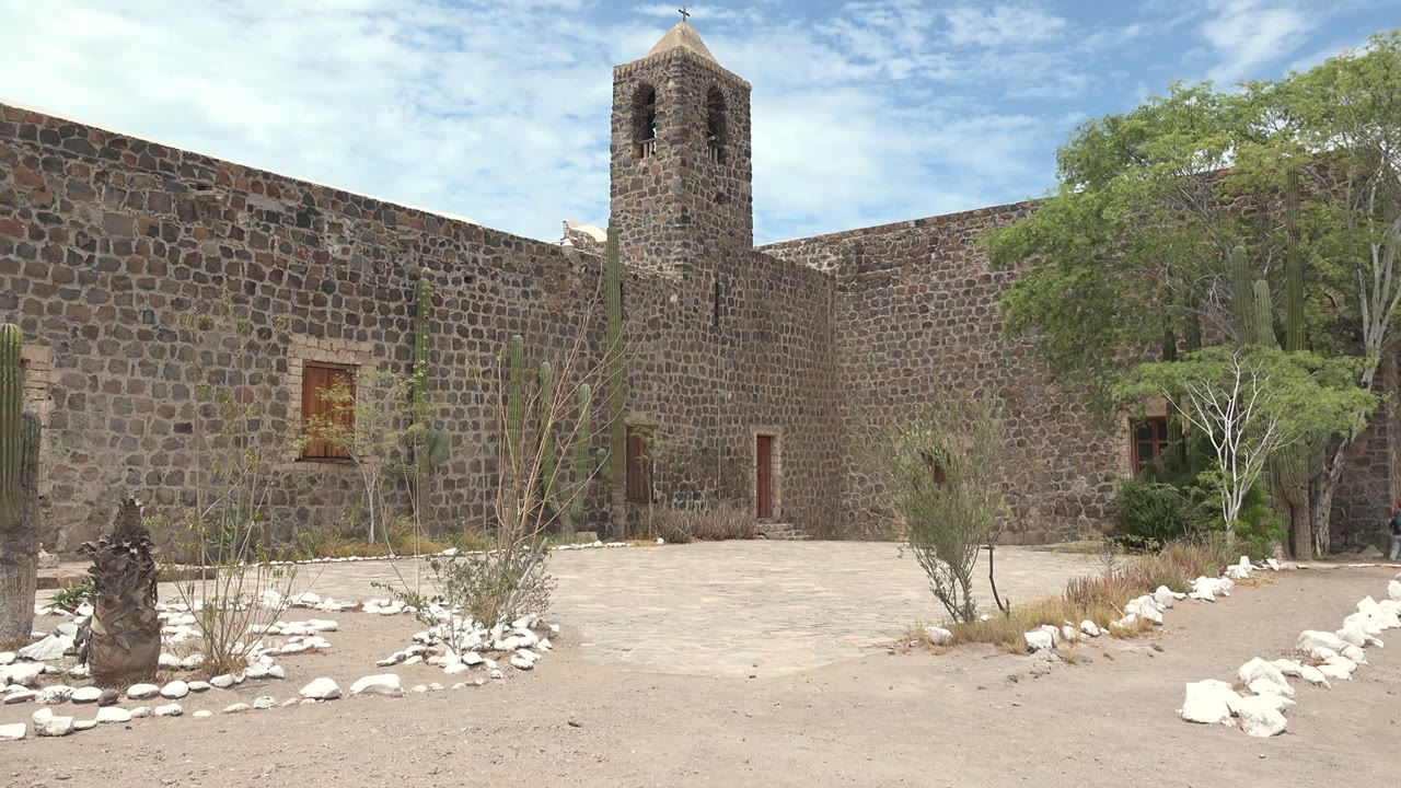 Stone bell tower and mission wall behind cacti and desert landscaping.