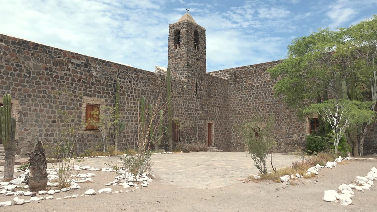 The Mulege courtyard remains empty as the stone bell tower, rough masonry wall, foreground cactus, trees, pale ground, and patchy clouds stay in view with no visible people.