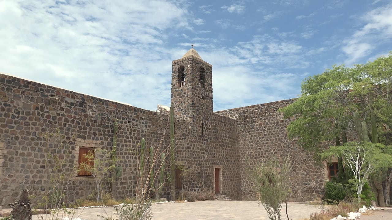 Mulege, Baja California Sur stone mission courtyard with the bell tower left of center, cacti and dry shrubs in front, a green tree to the right, and no visible people.