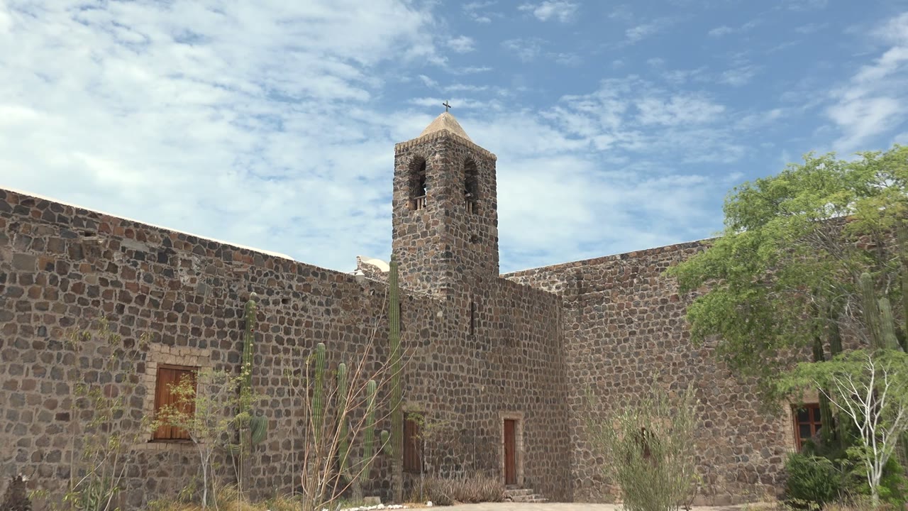 Stone bell tower and mission wall with desert plants under scattered clouds.