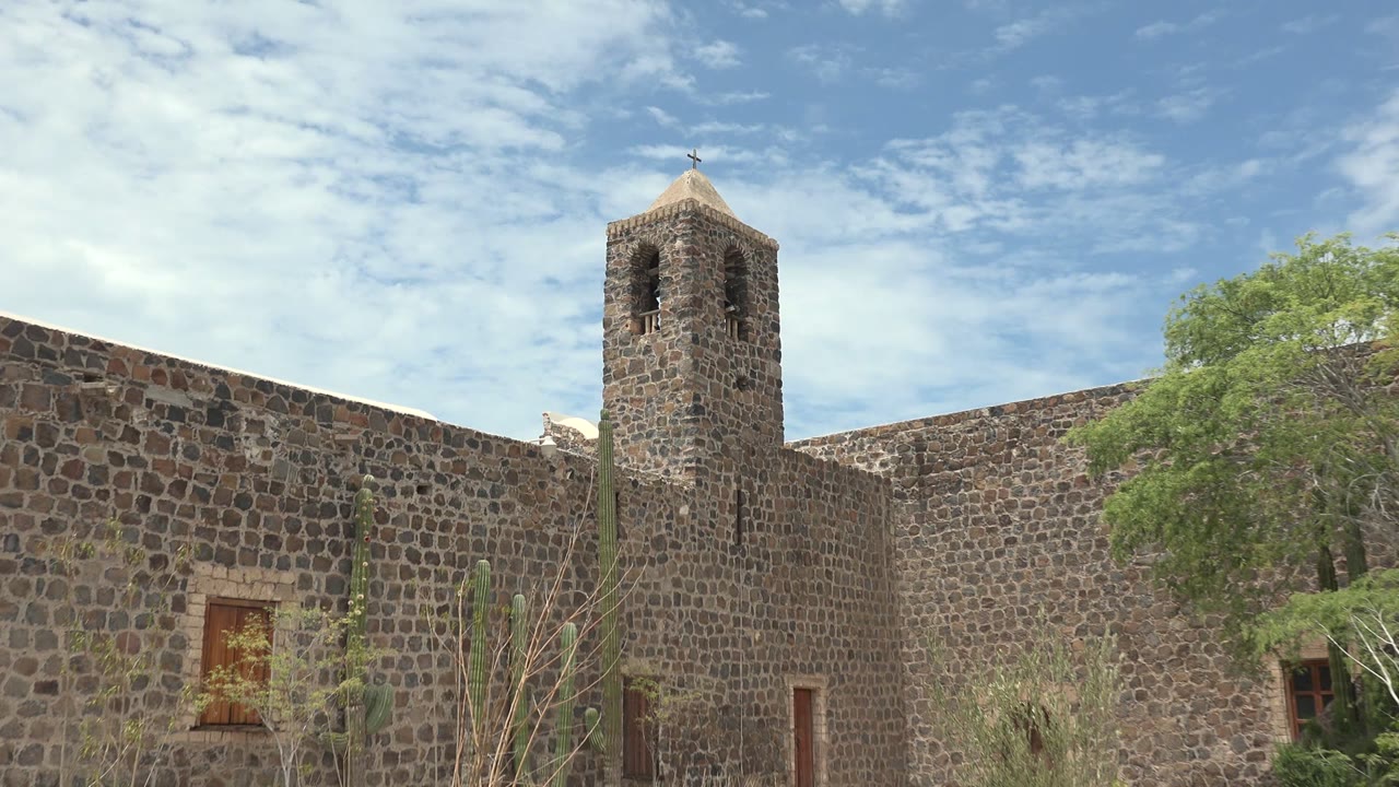Mulege, Baja California Sur stone mission wall with the bell tower rising above the courtyard, cacti and thin branches in front, green foliage at right, and no visible people.