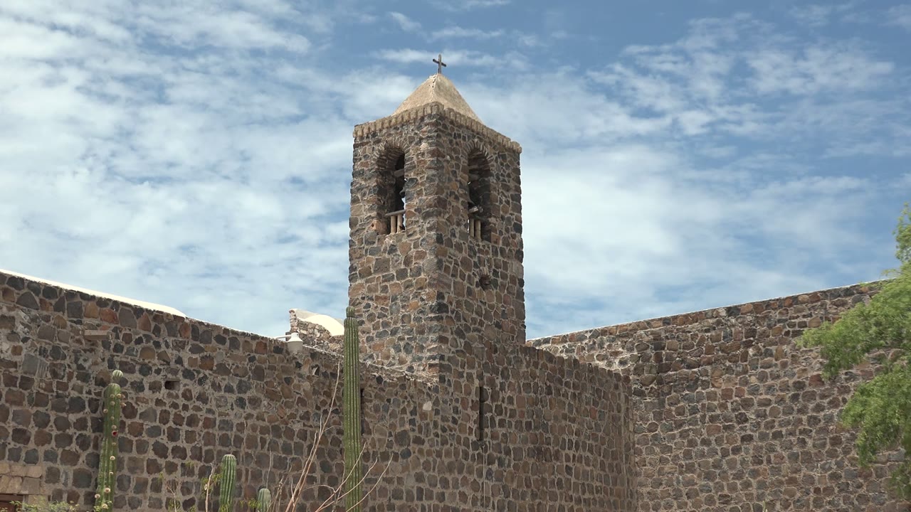 At 0:00 in Mulege, Baja California Sur, a stone church tower with a cross rises behind a stone wall, with cacti and a small tree visible under scattered clouds, no visible people.