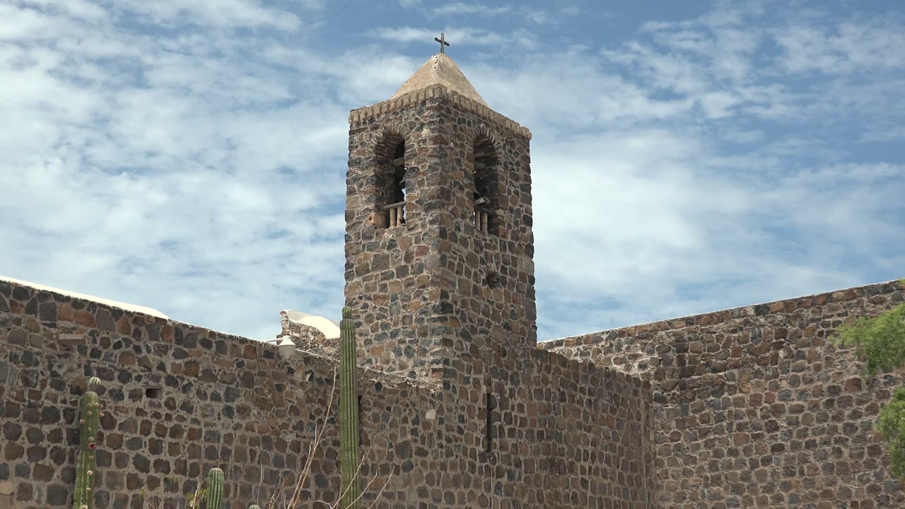Stone bell tower above a stone wall with cacti and scattered clouds.