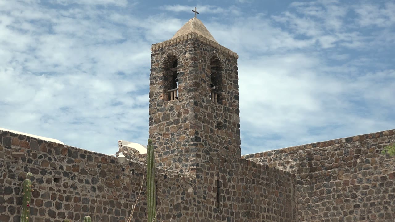 At 0:02 in Mulege, Baja California Sur, the tower remains high in frame with its dark bell openings visible, while the stone wall and cacti run across the lower edge, no visible people.
