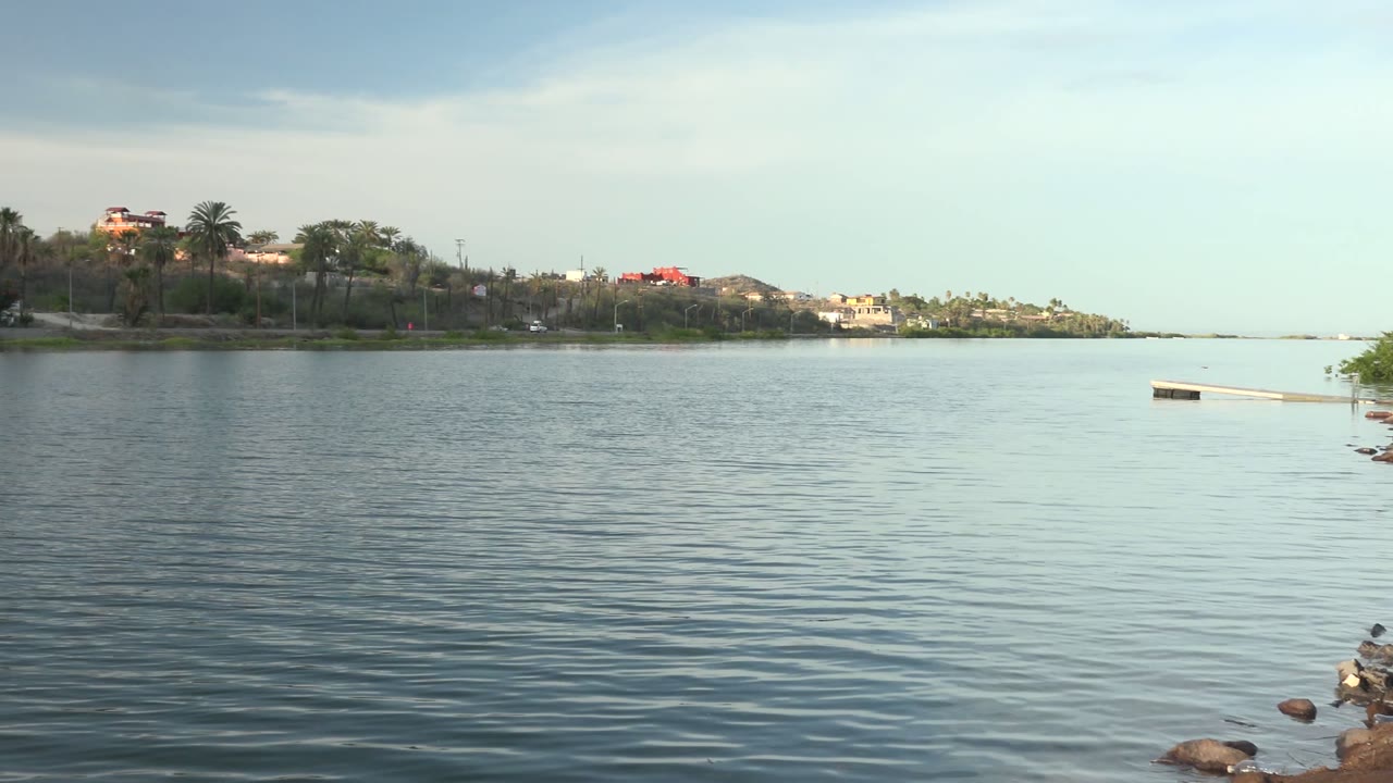Opening Mulege, Baja California Sur frame showing calm rippled water, distant trees and buildings across the water, a pale cloudy sky, and no visible people.