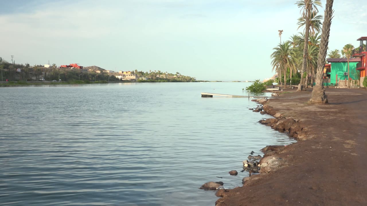 Opening frame of a Mulege, Baja California Sur waterfront, with rippled water on the left, a rocky sandy shore on the right, palms and buildings beyond, and no visible people.