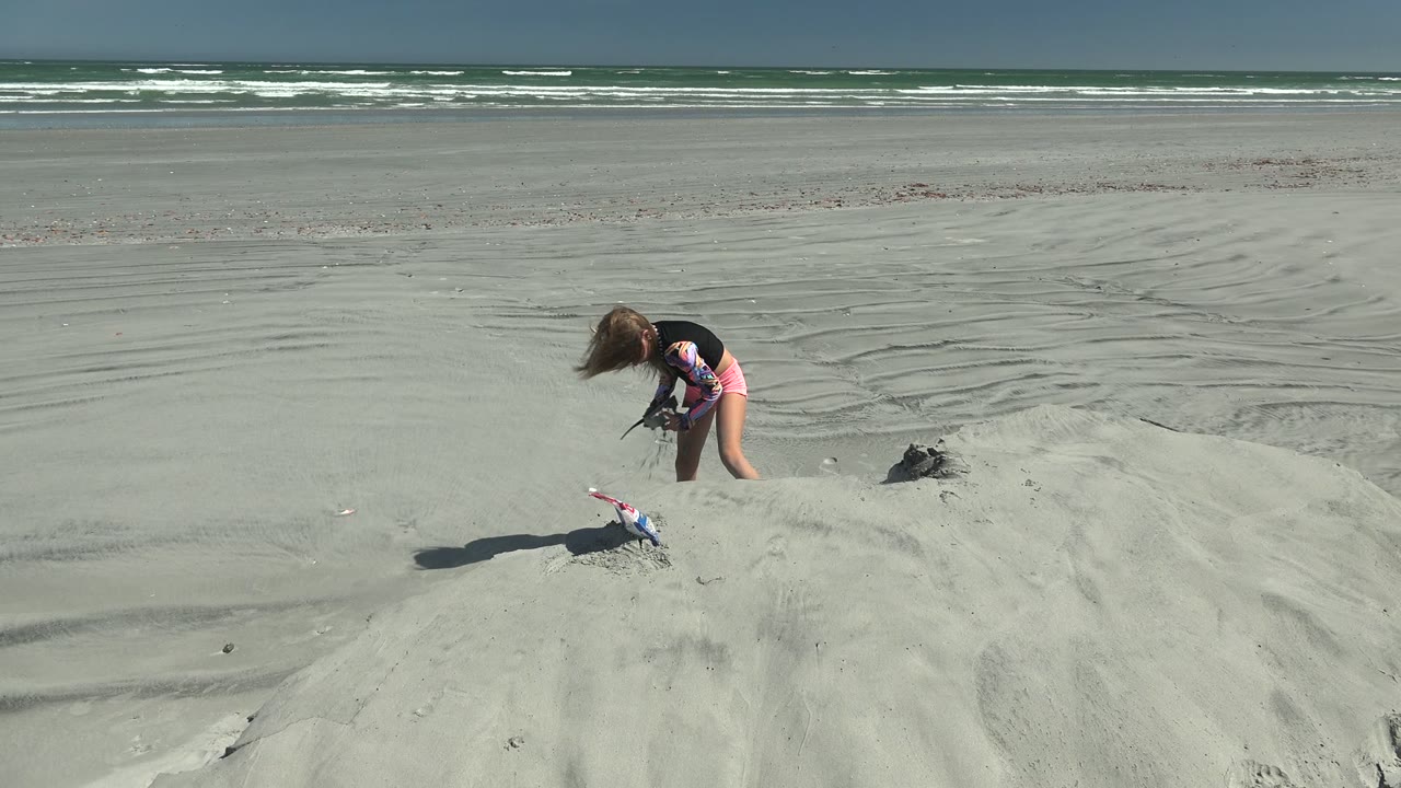 At 0:00 in La Bocana, Mexico, a young girl bends over a pale sand mound with a small shovel, a toy nearby, and ocean waves across the background.