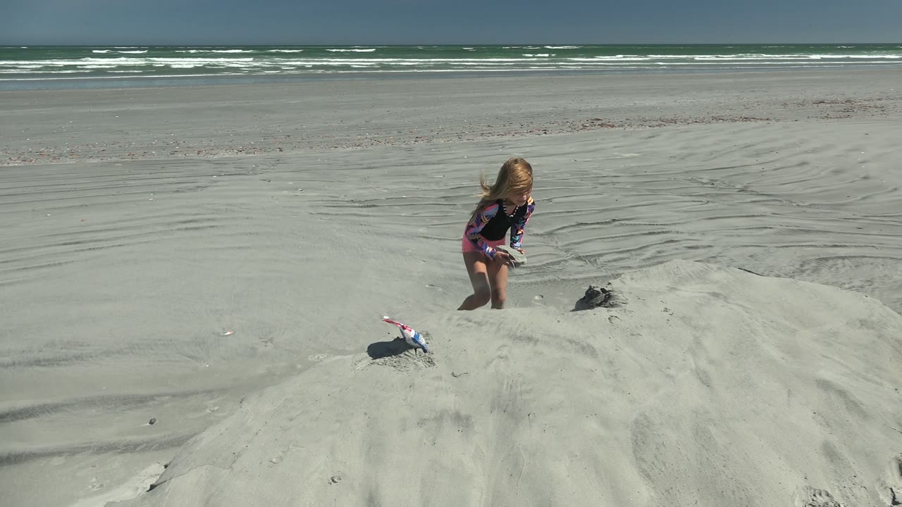 Young girl stands on the sand mound beside a toy while waves roll in the distance.
