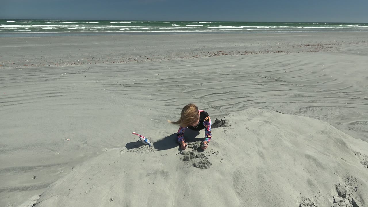 At 0:02 in La Bocana, Mexico, the girl kneels low on the sand mound, leaning into the sand with the toy still nearby and broad beach flats stretching to the surf.