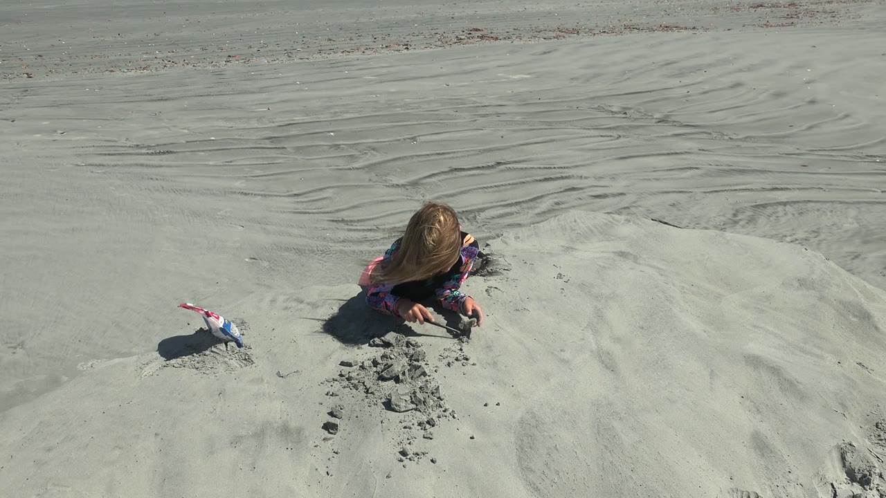 At 0:00 in La Bocana, Baja California Sur, a young girl lies low on pale sand and digs with her hands while a small toy shovel rests to her left.