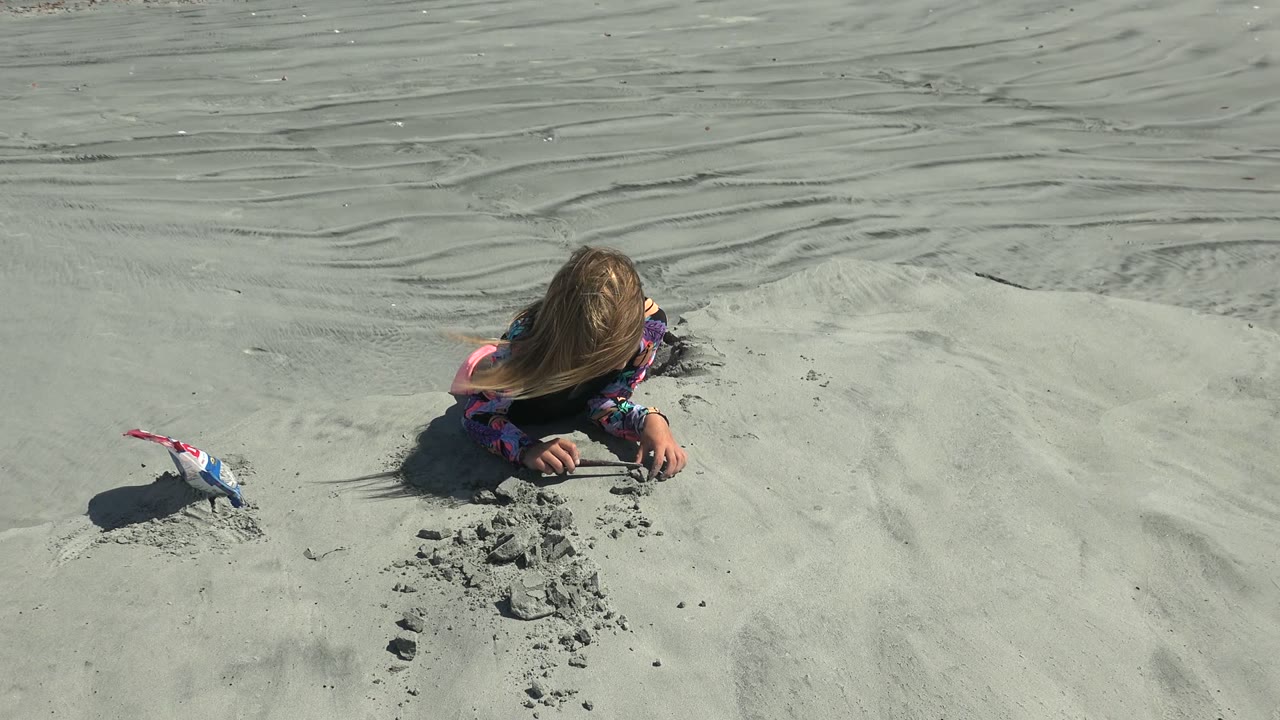 At 0:02 in La Bocana, Baja California Sur, the child digs into a small mound of sand with both hands while the toy shovel remains on the left side of the sandy frame.