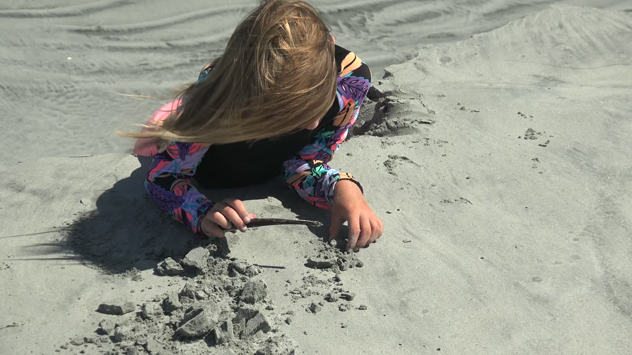 A long-haired child in a colorful shirt leans over pale sand in La Bocana, Baja California Sur, using a stick near small sand piles.