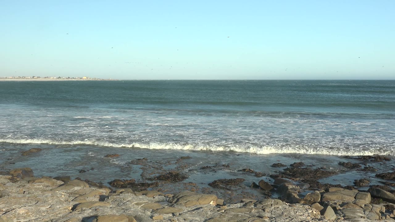 At La Bocana, Baja California Sur, ocean waves approach a rocky foreground under clear blue sky, with distant land on the horizon and no visible people.