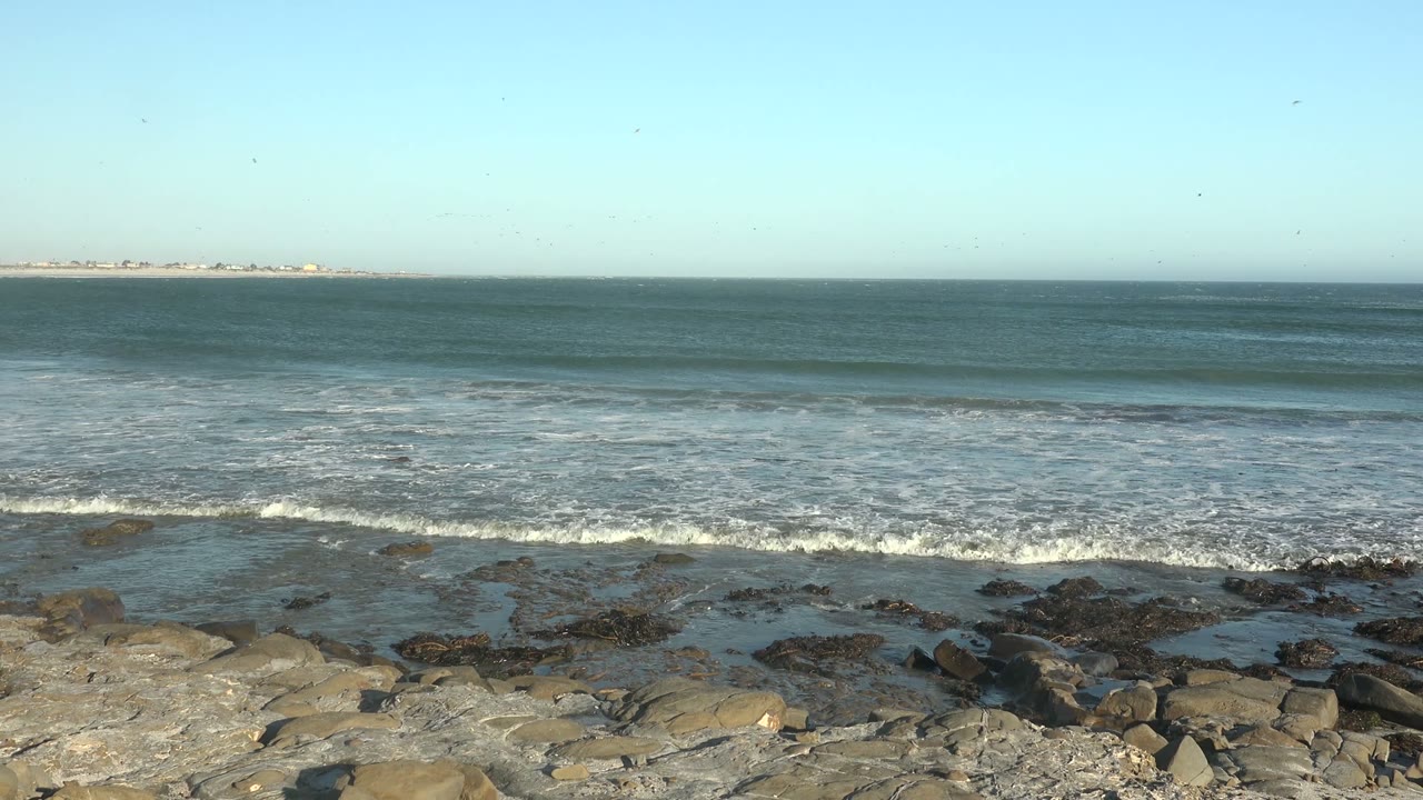 White surf breaks across rocks below a wide ocean horizon.