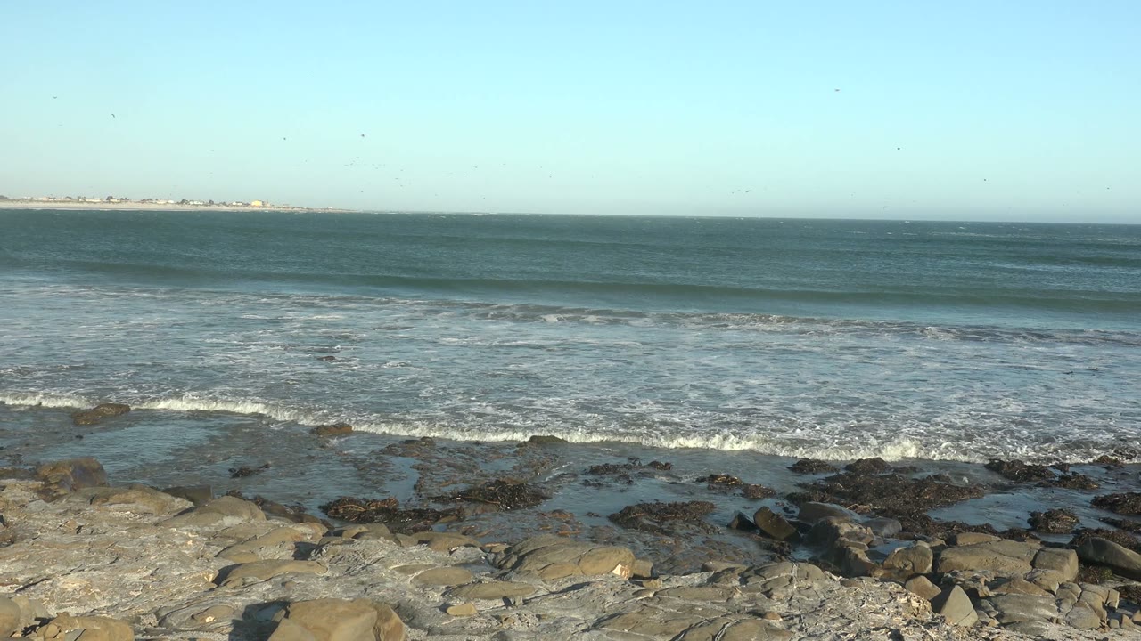 At La Bocana, Baja California Sur, low ocean swells roll toward the rocky waterline as foam gathers along the shore, with no visible people.