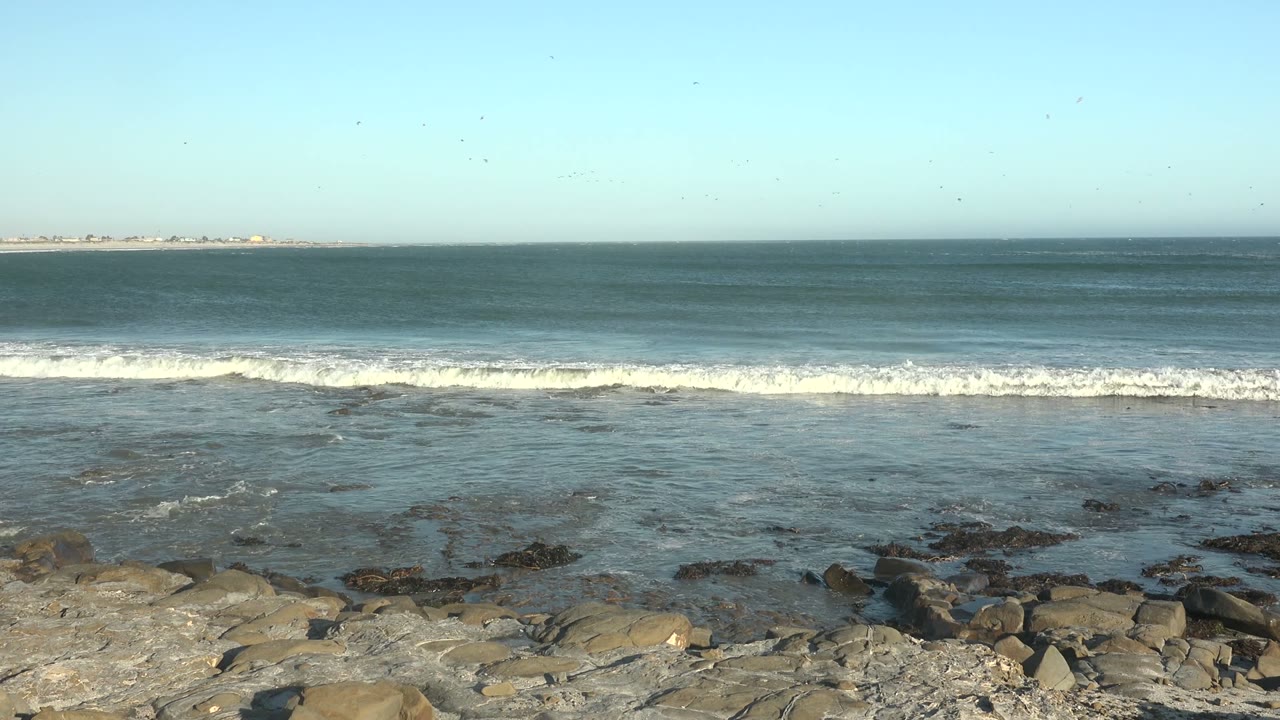At 0:00 in La Bocana, Baja California Sur, white surf lines approach a rocky foreground under a clear pale blue sky, with no visible people.