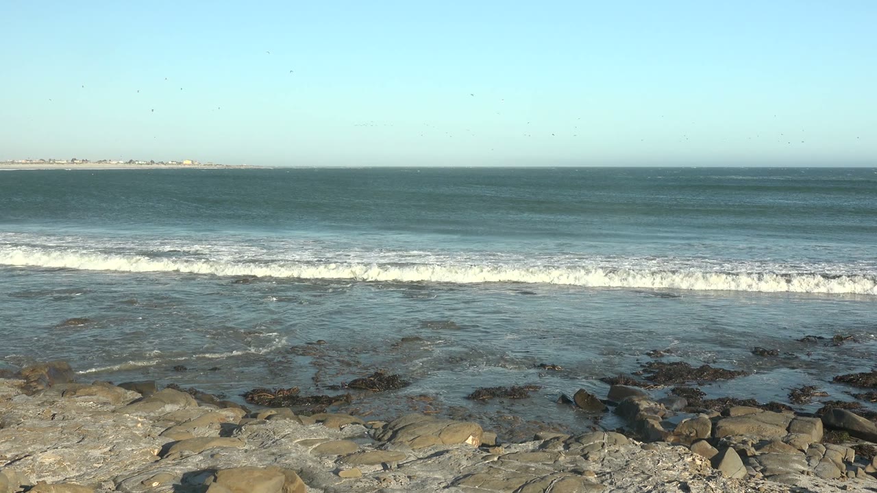 At 0:02 in La Bocana, Baja California Sur, the foam band spreads wider across the water while rocky foreground texture remains visible, with no visible people.