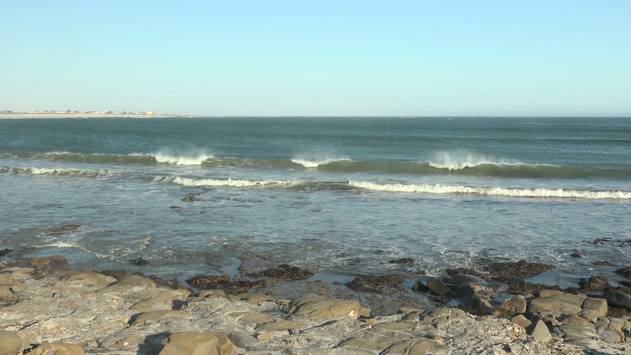 At La Bocana, Baja California Sur, blue ocean waves roll toward a rocky foreground with a faint landmass on the horizon and no visible people.