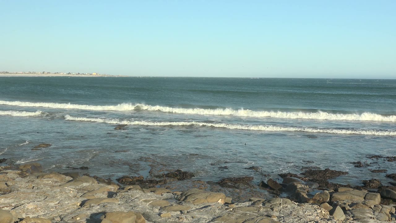 At La Bocana, Baja California Sur, parallel surf bands move across the blue water toward foreground rocks, with the horizon held high and no visible people.