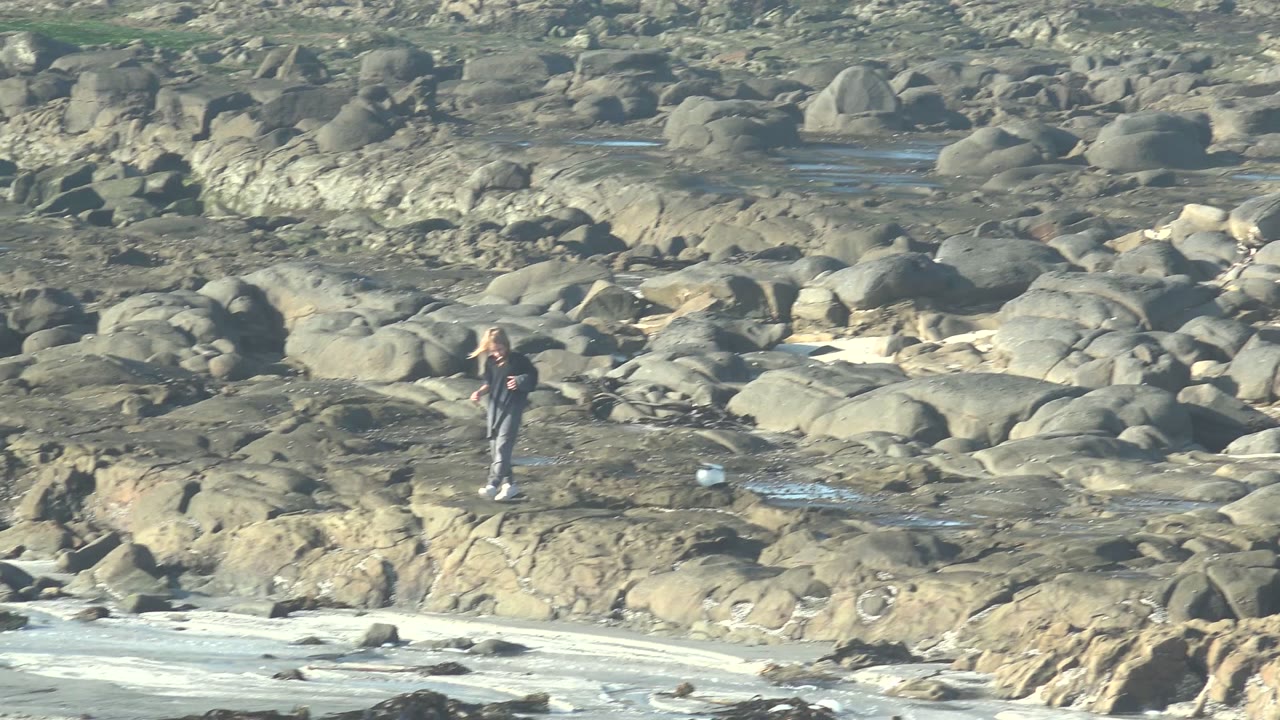 A person stands upright among rounded rocks in La Bocana, Mexico, with the white bucket set to one side and a band of foamy water crossing the foreground.