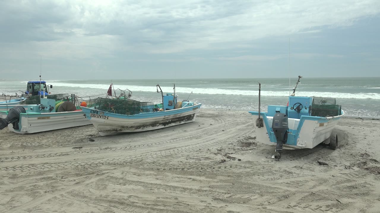 Small blue and white fishing boats rest on tracked sand at Punta Abreojos, Baja California Sur, with surf breaking behind them under a cloudy sky.