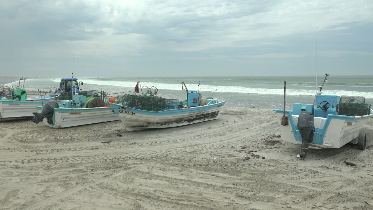 Several small boats remain parked on the sandy Punta Abreojos shoreline, including a foreground boat stern at right and surf stretching across the horizon.