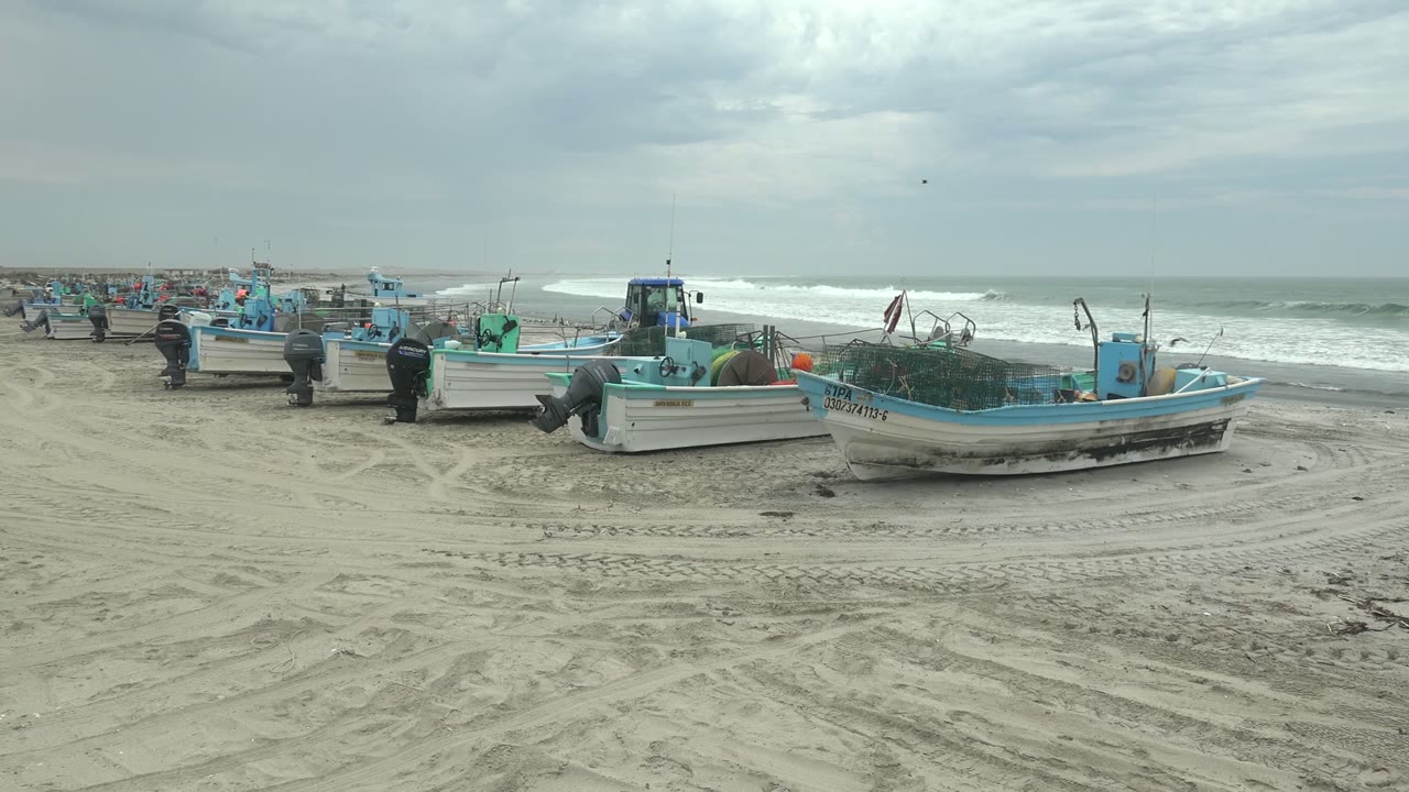 Fishing boats rest on tire-marked sand at Punta Abreojos, Baja California Sur, with surf and a cloudy horizon behind them and people visible near the boats.