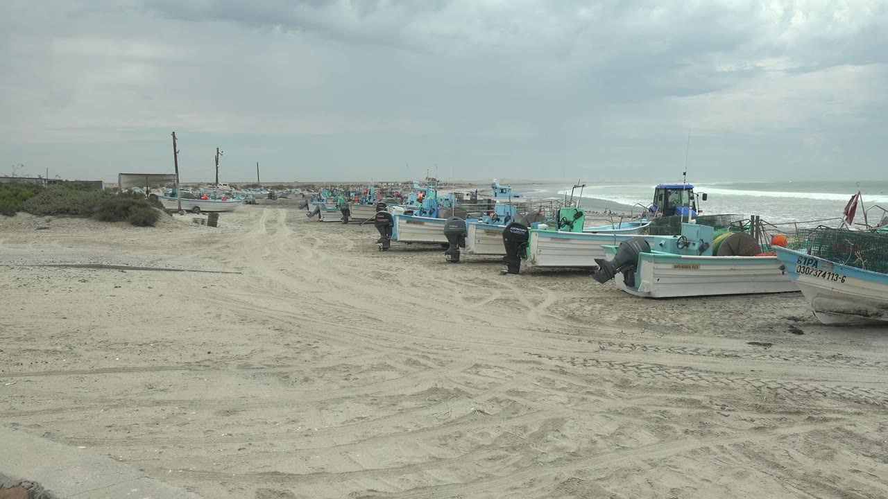 Opening view in Punta Abreojos shows fishing boats lined along tire-marked sand with ocean surf behind them and a few people near the vessels.