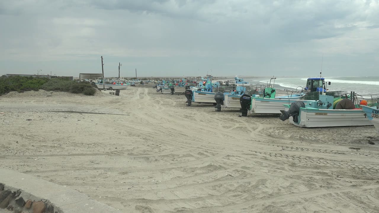 The Punta Abreojos beach remains framed wide, with several boats lined to the right, open sand through the center, low vegetation at left, and gray cloud cover above the surf.