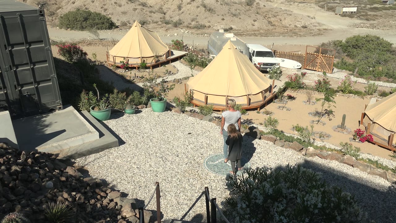 At 0:00, an adult and child walk across pale gravel in San Juanico, Baja California Sur, with tan yurts, potted plants, a white van, and dry hills beyond them.
