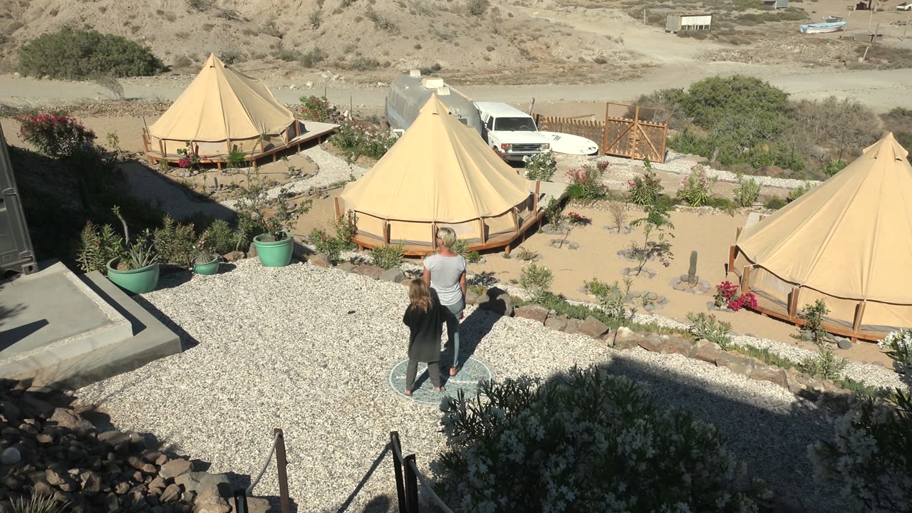 At 0:02, the walking pair appears nearer the center of the gravel area, with a large tan yurt to the right, another yurt to the left, and the white van parked behind the camp.